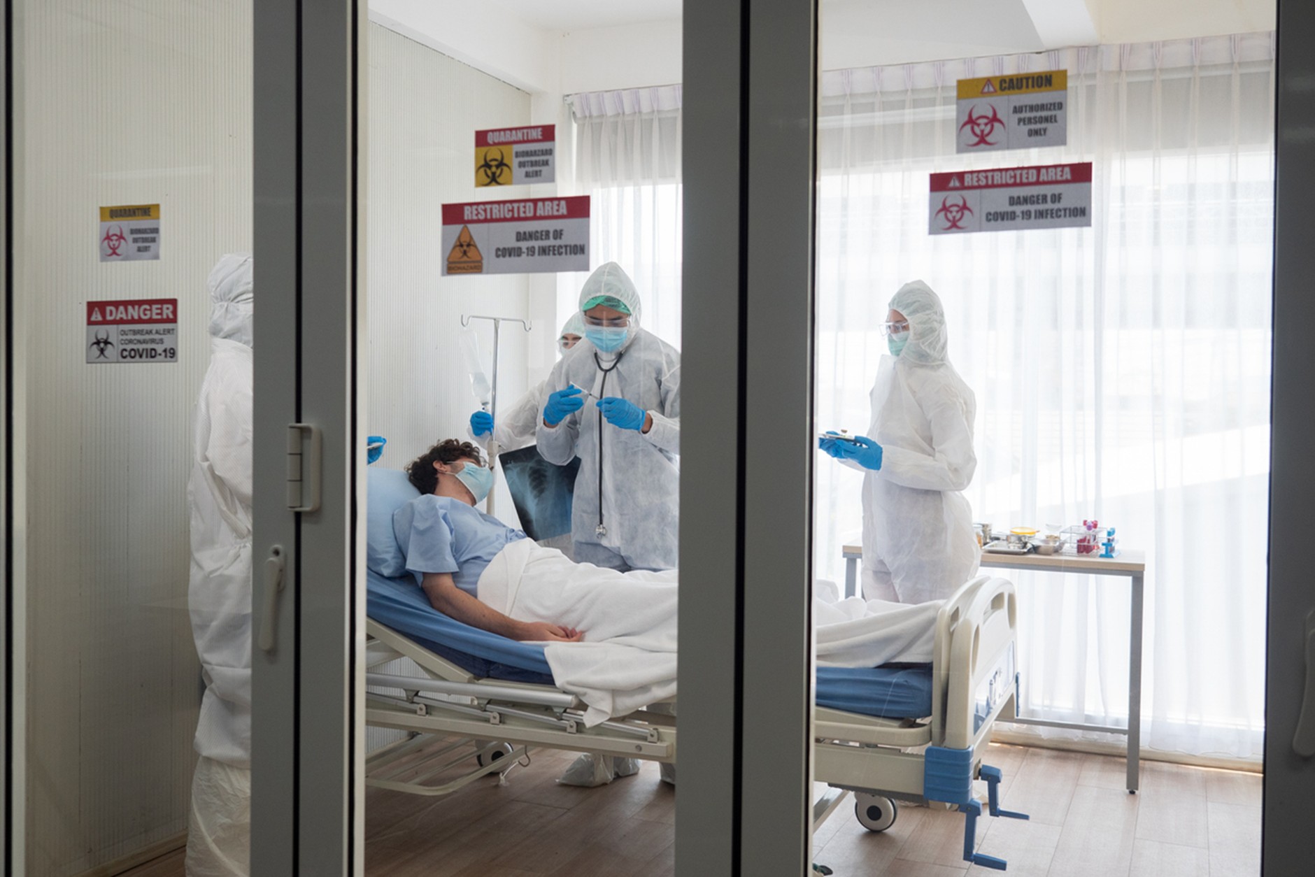 Healthcare workers wearing full protective equipment care for a patient in an isolation room marked with COVID-19 quarantine and restricted-area warning signs, viewed through glass doors.