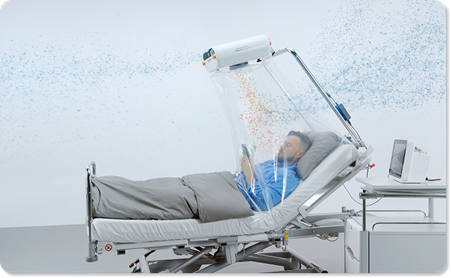 A person lies in a hospital bed under a transparent enclosure while a mounted device releases a fine mist or airflow above them; a medical monitor and equipment sit beside the bed in a clean clinical setting.
