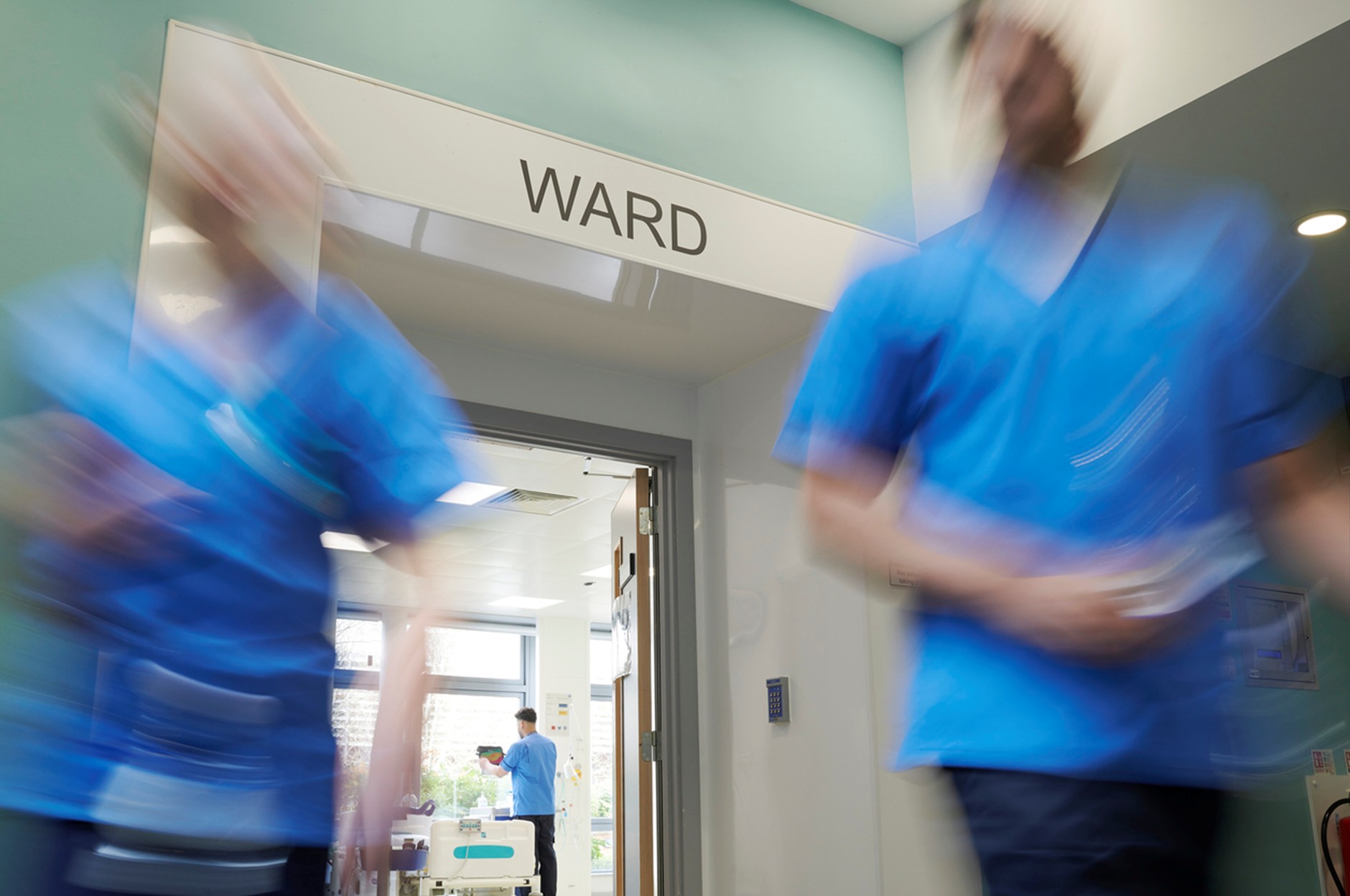 Blurred healthcare workers in blue scrubs move through a hospital ward entrance beneath a sign reading “WARD,” with a patient room visible in the background