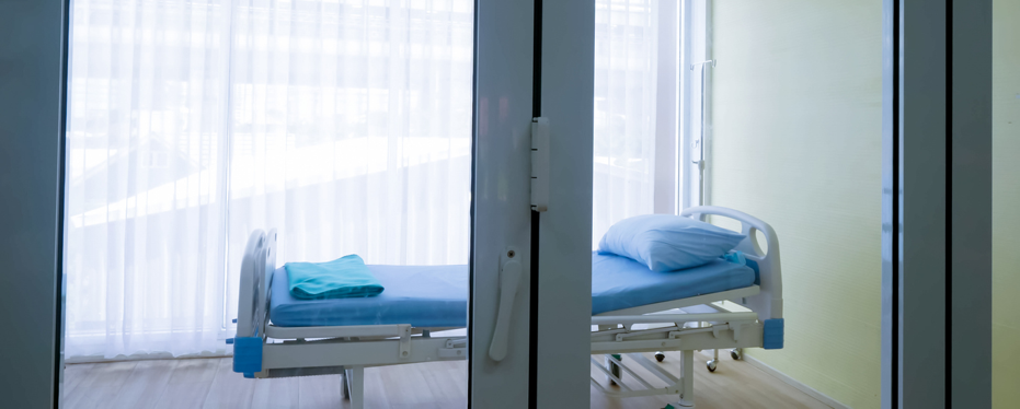Empty hospital patient room with a single adjustable bed, pillow and folded blanket, viewed through a glass door with natural light filtering through large curtained windows.