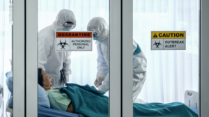 Patient lying in a hospital bed inside an isolation room while two healthcare workers in full protective suits provide care. Warning signs on the glass door read “Quarantine - Authorized Personnel Only” and “Caution - Outbreak Alert.”